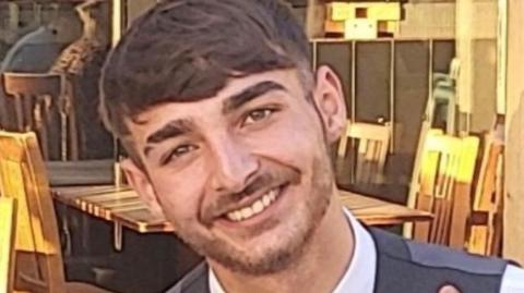 A young man with short dark hair and a short beard sits smiling at an outdoor table.