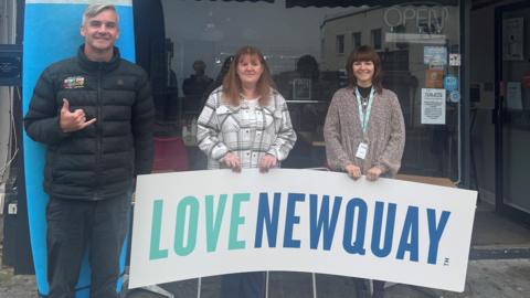 A man and two women are standing in front of a shop. The women hold a sign reading LOVE NEWQUAY while the man is standing in front of a paddle board making a hang-five surfer hand signal.