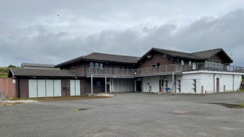 A disused building, previously used as a sports centre. Shutters are down around the building, and grey skies above it. 