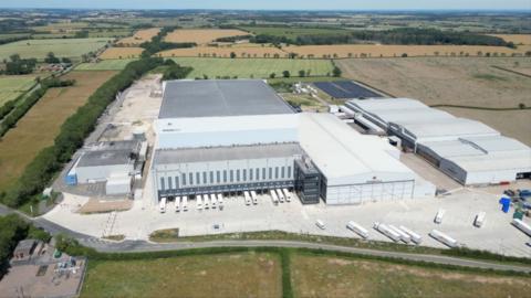 An aerial view shows a large industrial warehouse complex with multiple loading bays and lorries, surrounded by expansive farmland and fields.