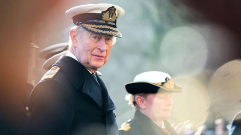Dressed in navy blue uniform King Charles III takes the salute from the newly commissioned naval officers on the march past during the Lord High Admiral's Divisions at Britannia Royal Naval College in Dartmouth. It is a sunny day and new officers also wearing navy uniforms can be seen in the background.
