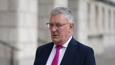 Mike Nesbitt has short grey hair and is wearing metal-framed glasses, a dark suit jacket, white shirt and pink tie