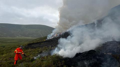 Cuideigin ann an aodach orains a' feuchainn ri smachd fhaighinn air teine fiadhaich.