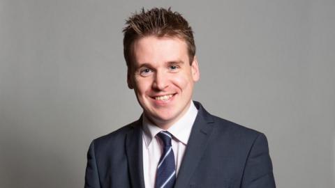 Tom Hunt is wearing a blue suit and is smiling at the camera. He's against a grey background and is wearing a blue and white striped tie. 