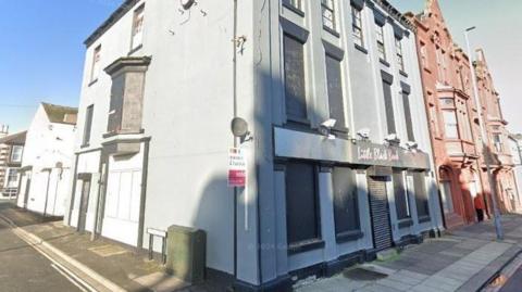 The exterior of the Little Black Book building. It is three storeys high with a shadow falling on the bottom half of the white building. Lots of windows have been boarded up.