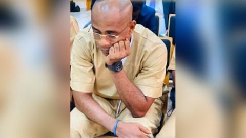 Baltasar Ebang Engonga, wearing glasses and dressed in beige prison clothes, sits with his head on his hand - leaning forward and looking down in court.