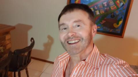 A man with short brown hair and a pink and white striped shirt in a room with a dark wooden chair to one side and a picture hanging on the wall behind him