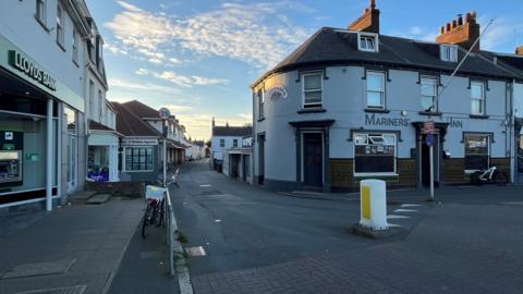 A quiet village scene with Nocq Road glowing under soft afternoon light. Lloyds Bank is in the left corner with its stone facade, while the a pub, the Mariners’ Inn stands opposite. A 25mph (40.2kmh) sign marks the narrow road ahead, curving gently into the distance beneath a warm, partly cloudy sky.
