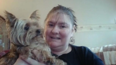 Ann Green is cuddling a pet dog and sitting on a sofa. She is wearing a black top and has her hair tied back in a ponytail and is wearing earrings.
