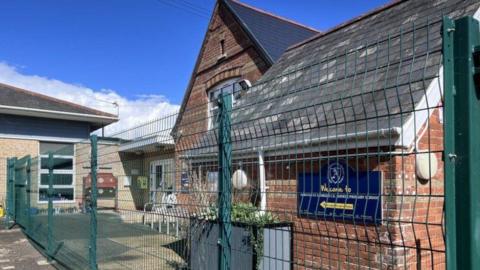 High green wire fence with brick building behind a navy blue sign, large planter and patio lead to a white door.