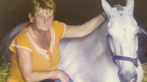 Eileen has short golden blonde hair and is wearing a yellow top. She is crouching next to a horse which is lying down in a bed of straw.