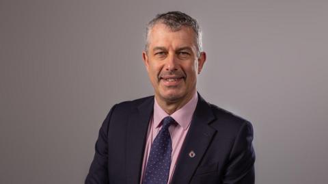 A picture of mark in a pink suit shirt, a blue tie and a black blazer.