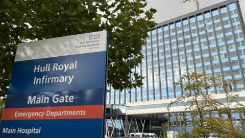 A multi-storey hospital building with blue cladding. A few trees and vehicles are in front of the building.