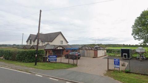 The image shows a building which appears to be a detached house and a driveway containing a black car and a trailer as well as a small boat on stilts. There are blue signs on a fence by the entrance to the driveway with white writing indicating the premises is Hatton Boarding Kennels.