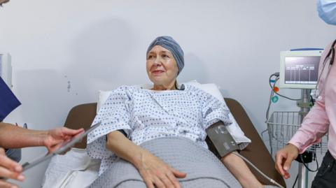 A stock image of a woman wearing a surgical gown and with a scarf over her head lying in a hospital bed with a blood pressure monitor on her left arm. Beside her is the screen the monitor is attached to and a man can be seen partly in the shot wearing a pink shirt and with a stethoscope around his neck. Only his arm and part of his chest can be seen.