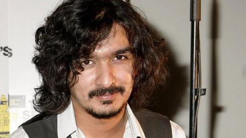 Boyan Chowdhury, who left The Zutons in 2007, poses for a photograph wearing a waistcoat and white shirt. He has long, curly black hair and a goatee beard.