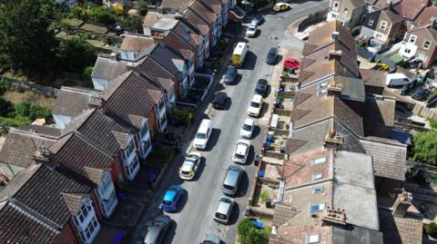 Two rows of terraces houses seen from above with vehicles, including police cars, parked on the street.
