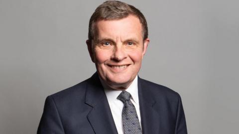 Profile photo of David Jones looking into camera wearing a dark suit, white shirt and grey tie, taken during his time in parliament.