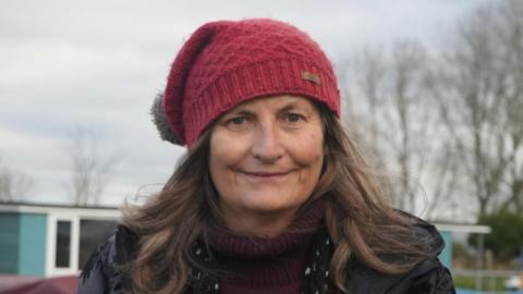 Woman stands on jetty and is wearing a red bobble hat, dark coat and red jumper. She has a black scarf with stars on it. Behind her are an array of boats.