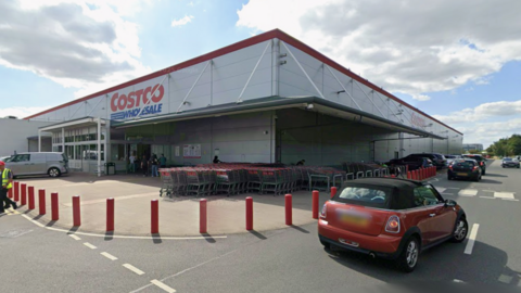 The Costco warehouse with blue sky behind and a red car at the front of the image