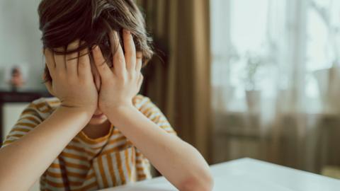 Anonymous boy with his head in his hands, elbows on a kitchen table, wearing a orange and white striped top.