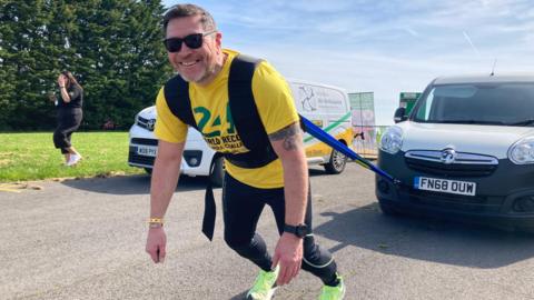 A picture of Mike Land, wearing a bright yellow t-shirt and a black harness, pulling a white van while smiling at the camera. 