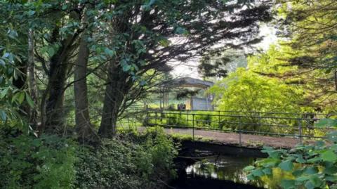 Part of the area, with a bridge over a river, trees, sun shining through leaves and part of an old barn in the background.