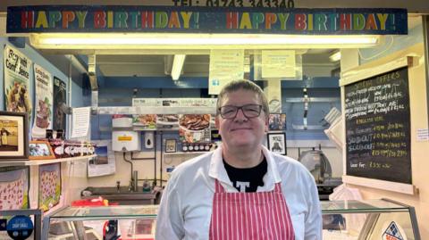 A man with brown hair and glasses is standing in front of a butcher's market stall. He is wearing a white shirt and red and white striped apron. There is a banner on the stall that reads "happy birthday!"