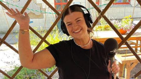 Jessie sat outside in a garden recording audio with a microphone in her hand and headphones on. Her arm is in the air and she's smiling. She is wearing a black t-shirt and pearl necklace.