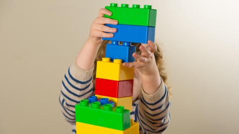 A generic image of a toddler who using large brightly coloured bricks to build a tower. The child is wearing a beige jumper with blue stripes and has curly yellow hair, but is largely hidden by the brick tower. 