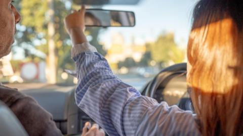 A woman sitting in the driver's seat of a car, adjusting her rear-view mirror with a male driving instructor sat next to her.