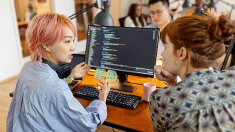 Stock photo shows a tech worker with pink hair sits at a desk and gestures while talking to a colleague who rests their chin on their hand, with other workers in the background, in an office environment with coding language on the computer screen in view.