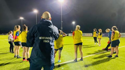 Image shows the a women's rugby team in a circle on the pitch, wearing yellow bibs. Neurodiversity coach Jacob Kelly looks on, wearing a navy jacket with his title on the back in white letters.