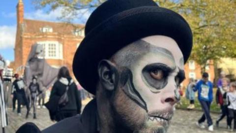 A man with skeleton face paint on and a green and black skeleton outfit side eye'ing the camera in the street. 