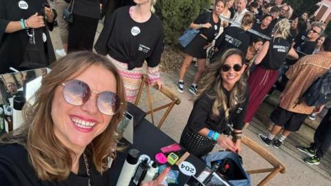 Anna and Claudia at the event. They are sat at a table which has varioud styling items on it like combs and hairspray. Both woman have long hair and sunglasses on and are smiling. They are wearing black shirts.  People are in the background queuing and watching on.