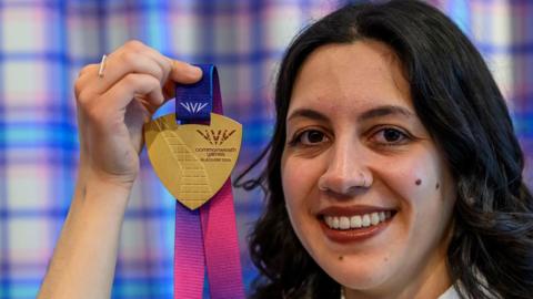 Militsa Milenkova - a woman with dark hair down to her shoulders, smiles while holding up a gold medal.