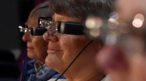 A close-up side view of three older women, all wearing black framed narrow smart glasses over their own spectacles 