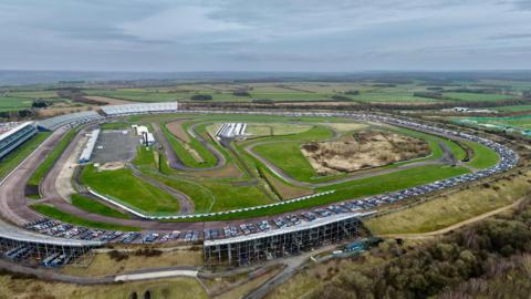 An aerial photo showing a large race track and grandstands surrounding the edge of the track. There is also a large car park. 