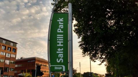 The sign for Salt Hill Park. It's on a post, with the backing in green and writing in white. It is dusk in the background.