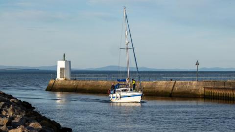 A small yacht sails through the entrance to Nairn Harbour. A white structure with a light to guide boats in is at the end of long wall at the entrance.