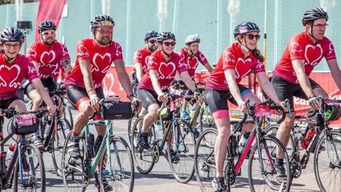A crowd of cyclists all wearing red jerseys with a heart on the front cycle on a road