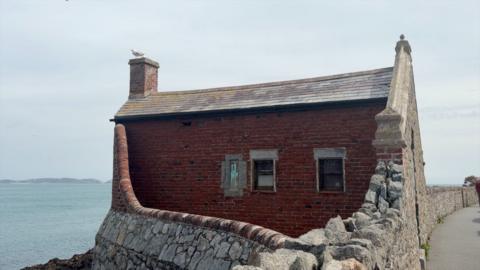 Photo of a one-storey red brick building with two small windows. There is a granite wall surrounding it.