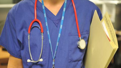 A medium shot of an unidentified doctor in uniform wearing an NHS lanyard and stethoscope around neck and carrying a file. 