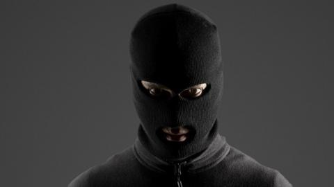 A man looks directly at the camera. He is wearing a black balaclava and a black zip-up top.