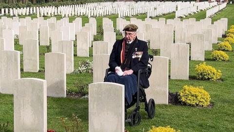 An elderly man wearing glasses and a military beret and blazer with rows of medals on his chest is sitting in a wheelchair among rows of headstones in a grassed cemetery. There are small yellow bushes at the end of each row.
