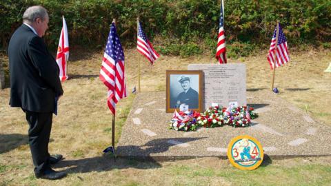 A picture of a man stood on grass. He is wearing a black suit with black shoes - facing a memorial. The memorial is to the right of where he is standing and has flowers and a picture in a gold frame on it. There are four American flags surrounding the memorial.