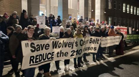 A large gathering of people in the dark, outside a large building with a white banner with the words Save Wolvey's Green Belt on it