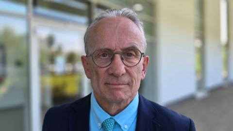 David Bick is wearing a dark blue suit jacket on top of a lighter blue shirt. He also has a blue patterned tie.