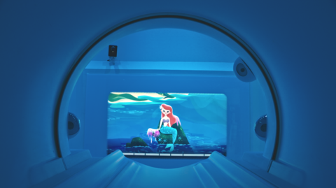 A view from inside an MRI scanner, looking through an archway with a cartoon image of a mermaid with red hair and a blue tail sitting on a rock in the sea at the end of the room. The room is lit in blue light.