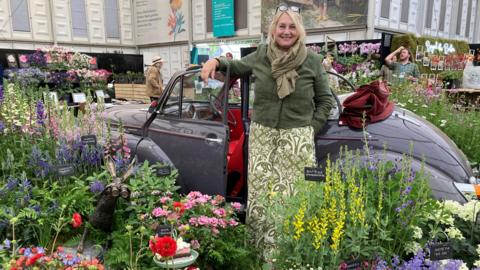 Stella Exley wearing a green jacket and a green and white long skirt stands in front of a Morris Minor with plants surrounding it at the Chelsea Flower Show.